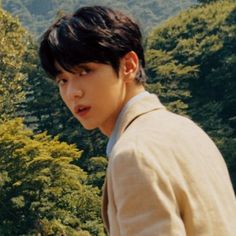 a young man standing in front of a forest with mountains and trees behind him, looking at the camera