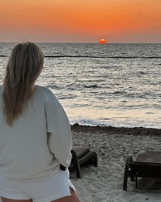 a woman is sitting on the beach watching the sun go down over the ocean with her legs in the air