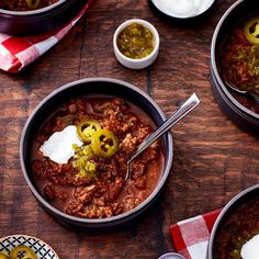 two bowls of chili with sour cream and peppers on the side next to spoons