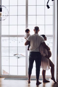 a man holding a baby in his arms while standing next to a window looking out at the water