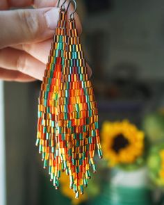 a person holding up a pair of multicolored beaded earrings with sunflowers in the background