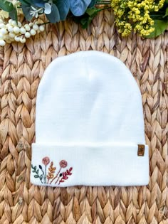 a white beanie with flowers on it sitting next to some green leaves and flowers