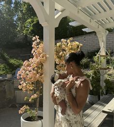 a woman holding a baby in her arms on a porch with flowers and trees behind her