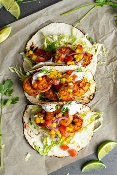 three tacos with shrimp, corn, and avocado on paper next to lime wedges