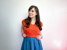 a woman standing in front of a white wall wearing an orange top and blue skirt