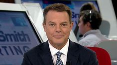 a man wearing a suit and tie sitting in front of a tv screen with another person behind him