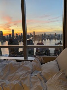 an unmade bed in front of a large window overlooking the city at sunset or dawn