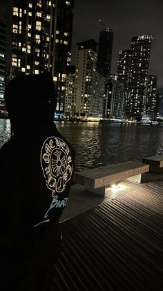 a person standing on a dock in front of the water at night with city lights behind them