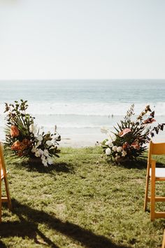 two wooden chairs sitting on top of a grass covered field next to the ocean with flowers in them