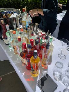 many bottles and glasses on a table with people in the background at an outdoor event