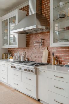 A bright white kitchen with a contrasting warm-toned brick backsplash. Red Brick Backsplash, Hood Ideas, Steel Range Hood, Stainless Range Hood, Stainless Steel Range