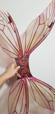 a person holding a butterfly shaped object in front of a white wall with red lines on it's wings