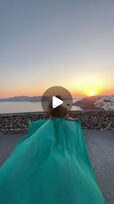 a woman in a blue dress sitting on a stone wall with the sun setting behind her