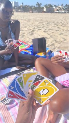 two women sitting on the beach playing cards with each other and one is holding a cell phone