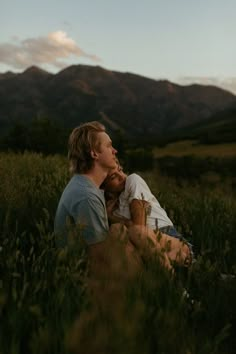 a man and woman sitting in tall grass with mountains in the background at sunset or dawn