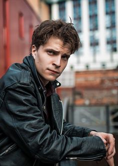 a young man leaning against a wall wearing a leather jacket