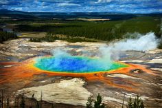 an aerial view of a geyser pool surrounded by trees and mountains in the distance