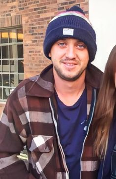 a man standing next to a woman in front of a brick building wearing a beanie