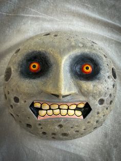 a creepy mask with red eyes and teeth on a gray cloth covered surface, that appears to have been painted