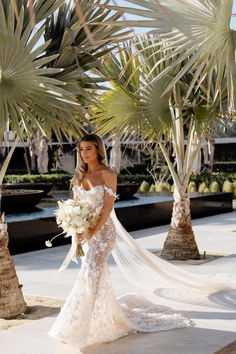 a woman in a wedding dress holding a bouquet and posing for the camera with palm trees behind her