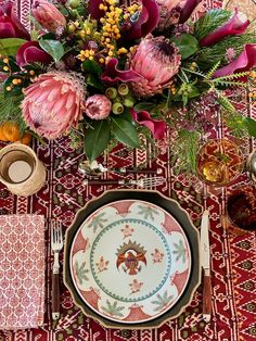 a place setting with flowers and plates on a red table cloth, next to an orange vase