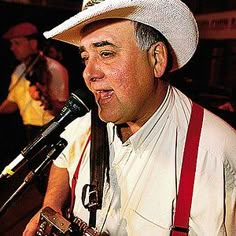 a man wearing a white hat and holding a camera in his right hand while singing into a microphone