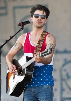 a man with sunglasses on playing an acoustic guitar and singing into a microphone while standing in front of a stage