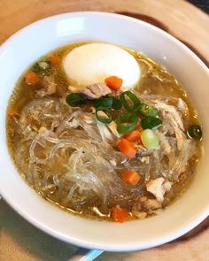 a white bowl filled with soup and vegetables on top of a wooden table next to a spoon