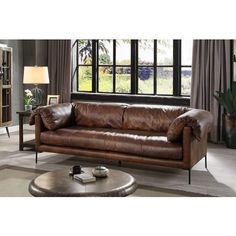 a brown leather couch sitting in front of a window next to a table and lamp