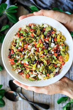 two hands holding a white bowl filled with colorful vegetables on top of a wooden table