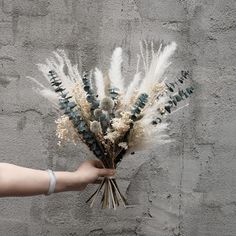 a hand holding a bouquet of dried flowers against a gray brick wall with writing on it