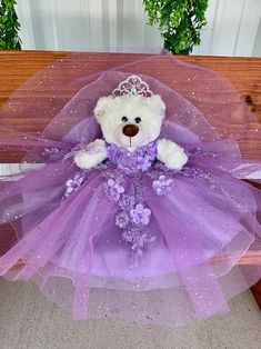 a teddy bear wearing a purple dress sitting on a bench with a tiara around its neck