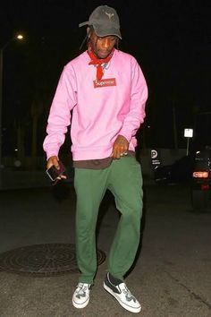 a man in a pink sweatshirt and green pants is standing on the street at night