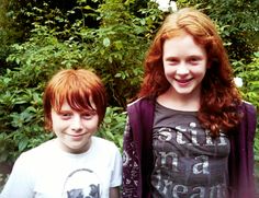 two young children standing next to each other in front of trees and bushes, smiling at the camera