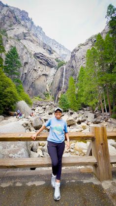 a woman standing next to a wooden bench in front of a mountain stream and waterfall