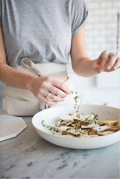 a woman sprinkling herbs on top of a white bowl filled with mushrooms in a kitchen