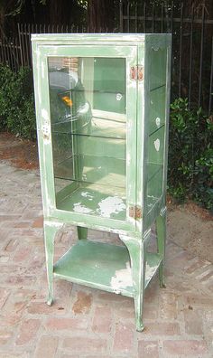 a green cabinet sitting on top of a brick floor next to a fence and bushes