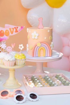 a table topped with two cakes and cupcakes next to balloons in the background