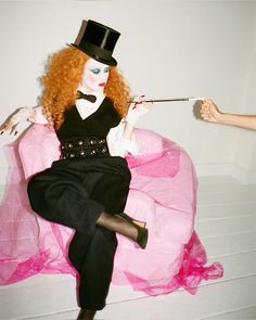 a woman with red hair wearing a top hat sitting on a pink chair and holding scissors