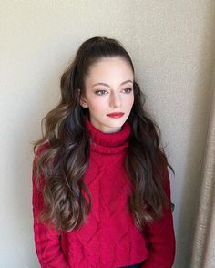 a woman with long hair wearing a red turtle neck sweater and black pants standing in front of a white wall