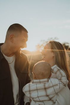 a man and woman holding a baby in their arms while the sun sets behind them