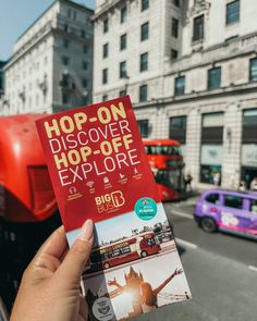 a person holding up a book in front of a red double decker bus on a city street