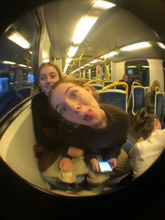 two women sitting on a train looking at their cell phones and taking a selfie