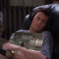 a man laying in a chair holding a book with headphones on his ears and eyes closed