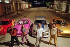 three men and two women standing in front of four different colored cars on the street