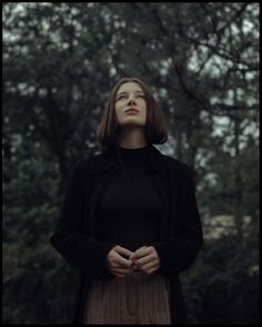 a woman standing in front of trees with her hands folded over her chest and looking up at the sky