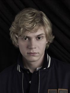 a young man with blonde hair wearing a black jacket and looking at the camera while standing in front of a dark background