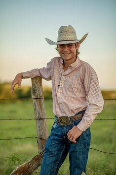 a man wearing a cowboy hat leaning against a fence in the grass with his hand on his hip