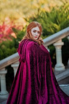 a woman with red hair wearing a purple cloak and posing in front of some stairs