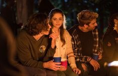 three people sitting next to each other in front of a campfire with one person holding a cup